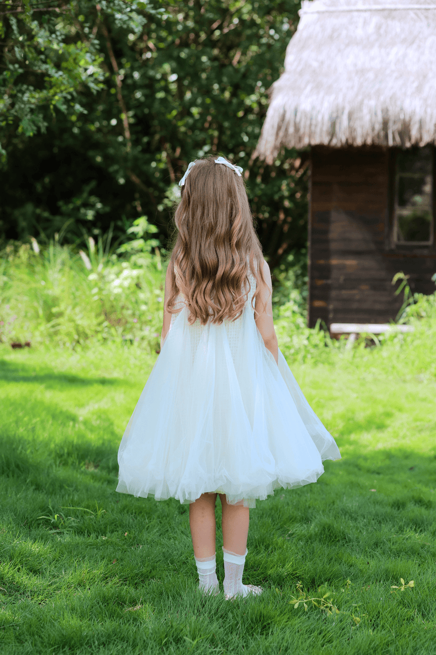 A young girl wears the KIKI and JOJO Girls Textured Velvet Sleeveless Embroidered Dress in Ivory, standing on green grass with a wooden hut and dense foliage in the background, facing away from the camera.