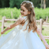 A young girl twirls outdoors near a wooden fence, wearing the KIKI and JOJO Girls Textured Velvet Sleeveless Embroidered Dress in Ivory, with greenery and trees in the background.