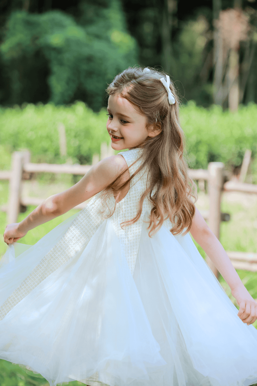 A young girl twirls outdoors near a wooden fence, wearing the KIKI and JOJO Girls Textured Velvet Sleeveless Embroidered Dress in Ivory, with greenery and trees in the background.