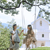 Two girls wearing the KIKI and JOJO Girls Velvet Asymmetrical Hem Puff Sleeve Dress in gray stand and sit on a swing hanging from a tree, with a white building in the background on a sunny day.