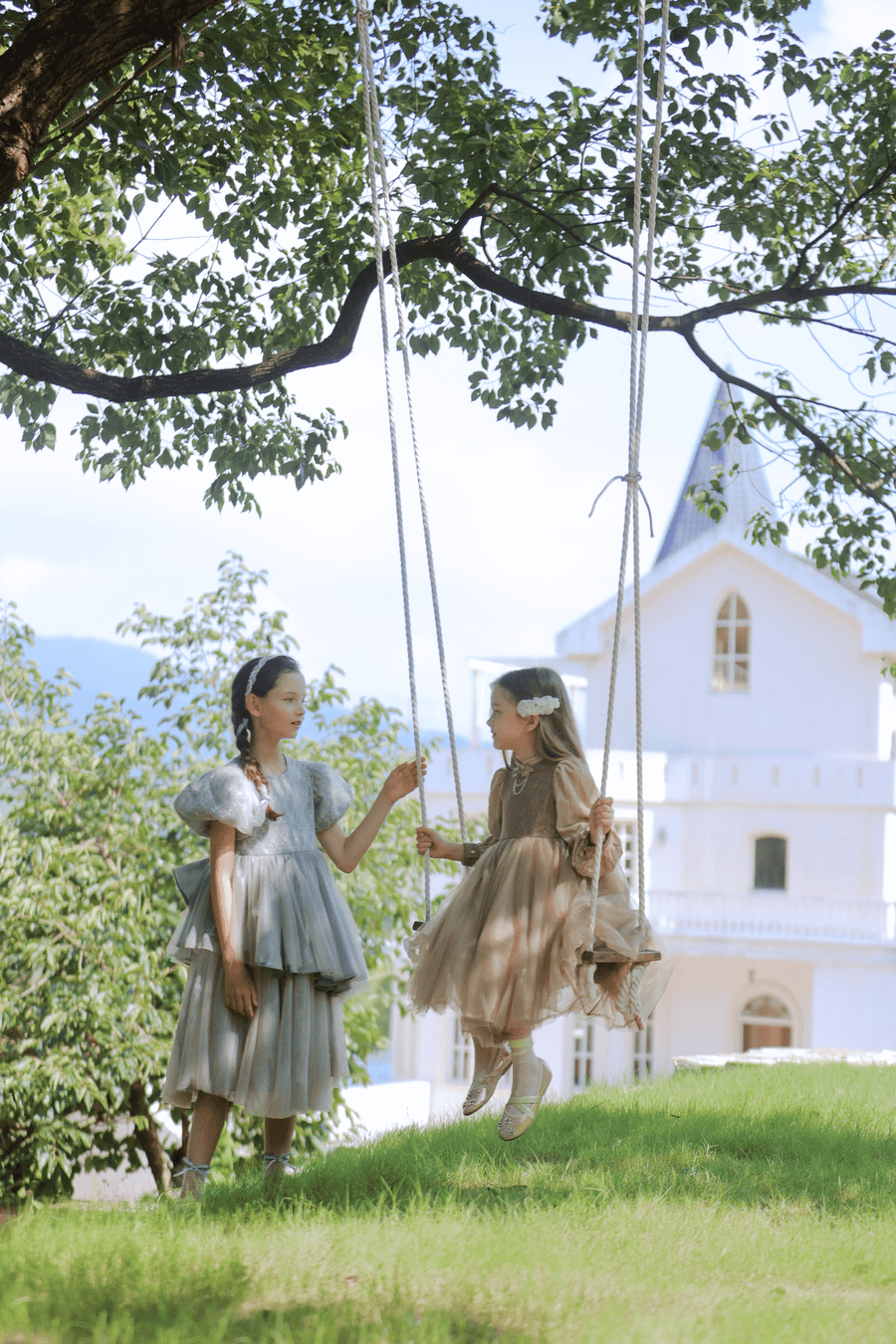 Two girls wearing the KIKI and JOJO Girls Velvet Asymmetrical Hem Puff Sleeve Dress in gray stand and sit on a swing hanging from a tree, with a white building in the background on a sunny day.