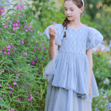 A young girl in a gray Girls Velvet Asymmetrical Hem Puff Sleeve Dress by KIKI and JOJO stands in a garden, holding a pink flower and admiring it, surrounded by greenery and more pink flowers.