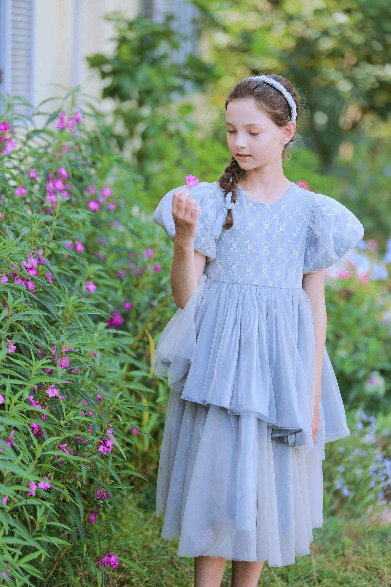 A young girl in a gray Girls Velvet Asymmetrical Hem Puff Sleeve Dress by KIKI and JOJO stands in a garden, holding a pink flower and admiring it, surrounded by greenery and more pink flowers.