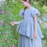 A young girl wears the KIKI and JOJO Girls Velvet Asymmetrical Hem Puff Sleeve Dress in gray, standing outdoors and gently touching pink flowers on tall green plants.