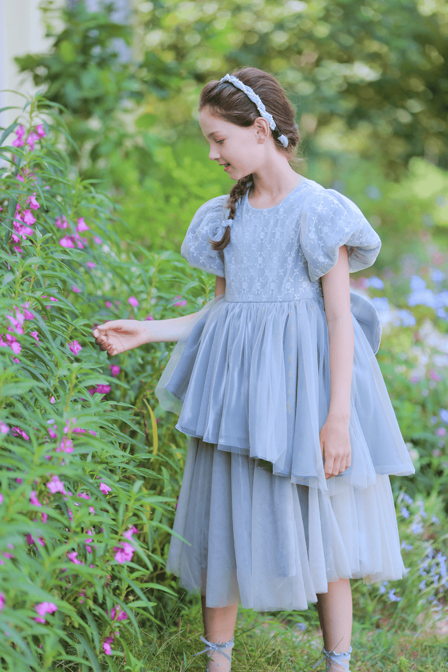 A young girl wears the KIKI and JOJO Girls Velvet Asymmetrical Hem Puff Sleeve Dress in gray, standing outdoors and gently touching pink flowers on tall green plants.