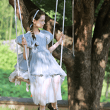 A young girl in a KIKI and JOJO Girls Velvet Asymmetrical Hem Puff Sleeve Dress in gray sits on a rope swing hanging from a tree, gazing to the side in a sunlit outdoor setting.