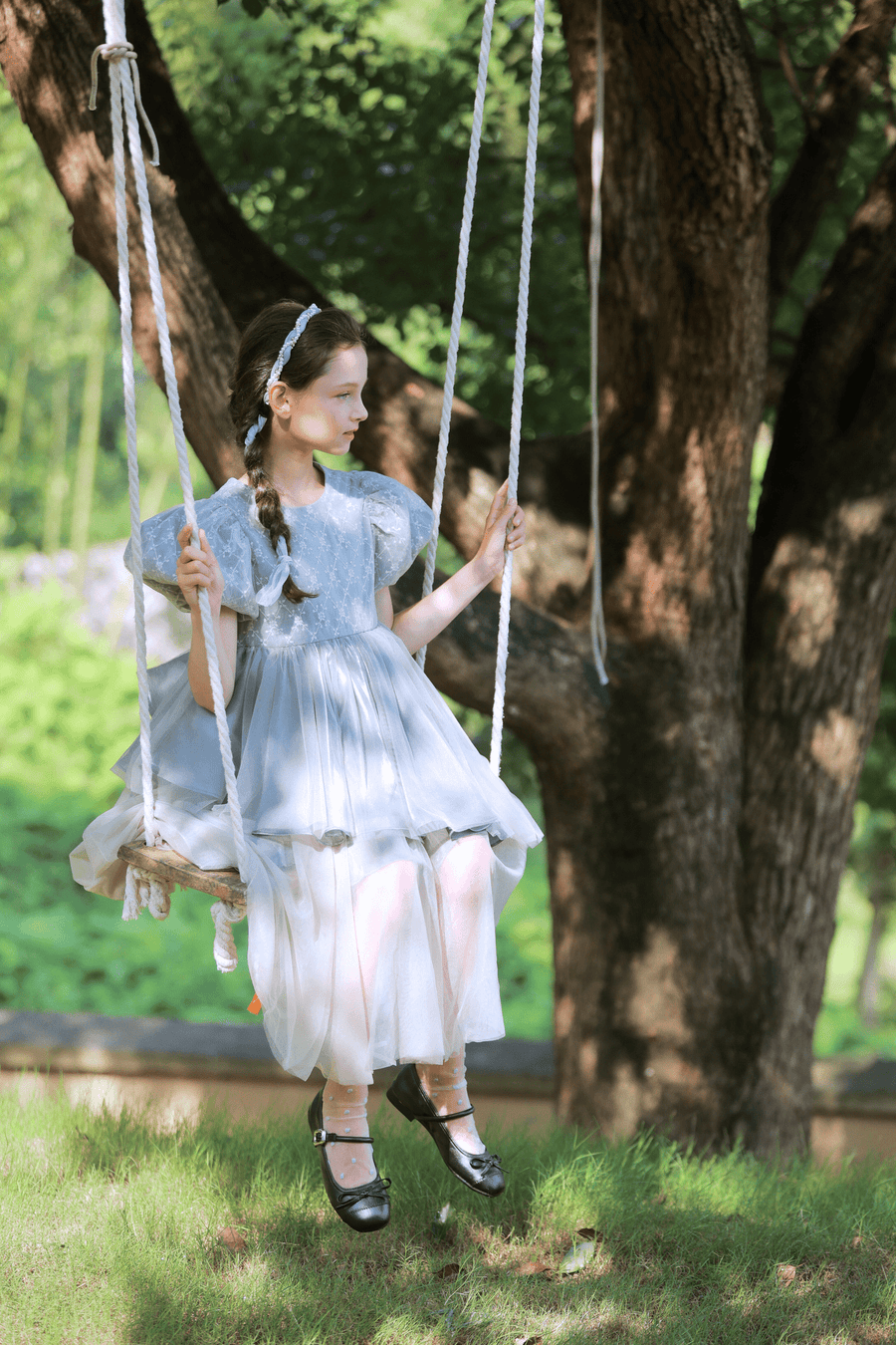 A young girl in a KIKI and JOJO Girls Velvet Asymmetrical Hem Puff Sleeve Dress in gray sits on a rope swing hanging from a tree, gazing to the side in a sunlit outdoor setting.