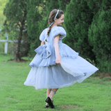 A young girl twirls on green grass near tall trees, wearing the KIKI and JOJO Girls Velvet Asymmetrical Hem Puff Sleeve Dress in gray.