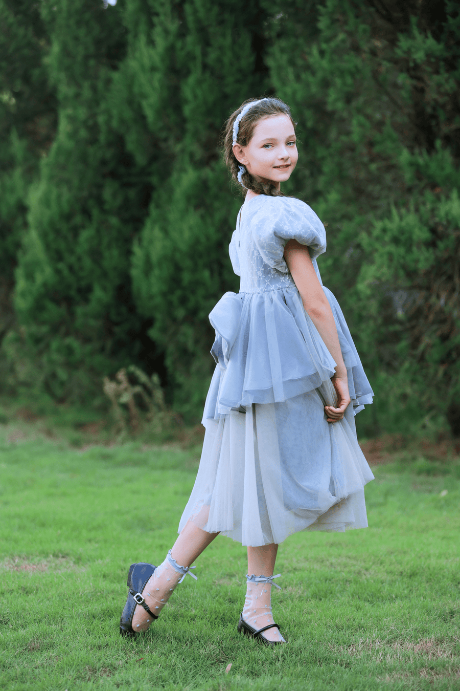 A young girl in a gray KIKI and JOJO Girls Velvet Asymmetrical Hem Puff Sleeve Dress with a headband stands on grass, looking over her shoulder, with green trees in the background.
