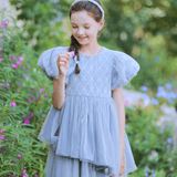 A young girl in a gray KIKI and JOJO Girls Velvet Asymmetrical Hem Puff Sleeve Dress stands outdoors, smiling with a small flower in hand, surrounded by greenery and pink flowers.