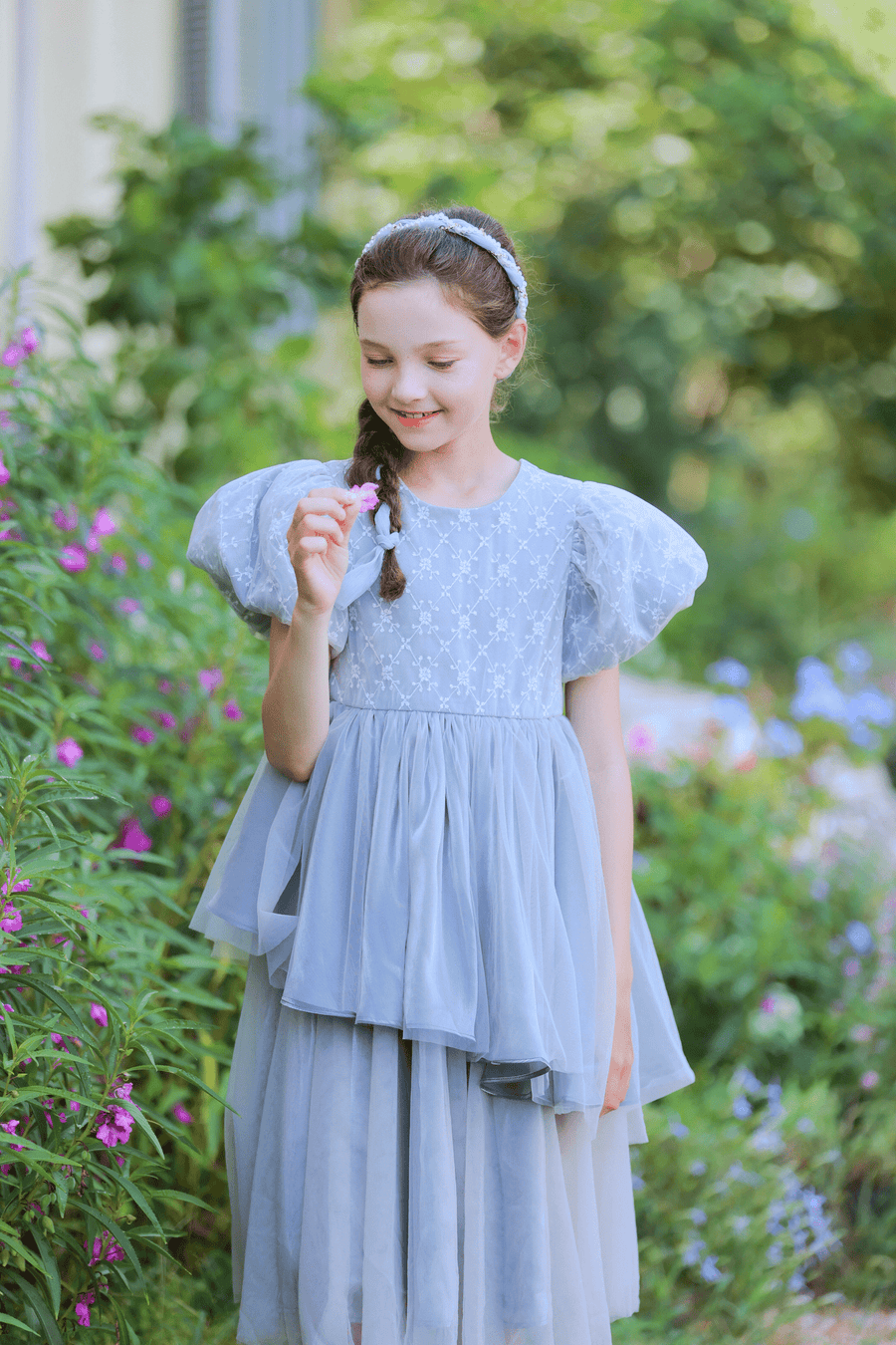 A young girl in a gray KIKI and JOJO Girls Velvet Asymmetrical Hem Puff Sleeve Dress stands outdoors, smiling with a small flower in hand, surrounded by greenery and pink flowers.