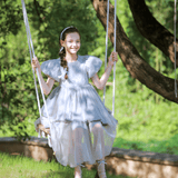 A young girl in the KIKI and JOJO Girls Velvet Asymmetrical Hem Puff Sleeve Dress in gray sits on a swing outdoors, smiling, with green trees and grass in the background.
