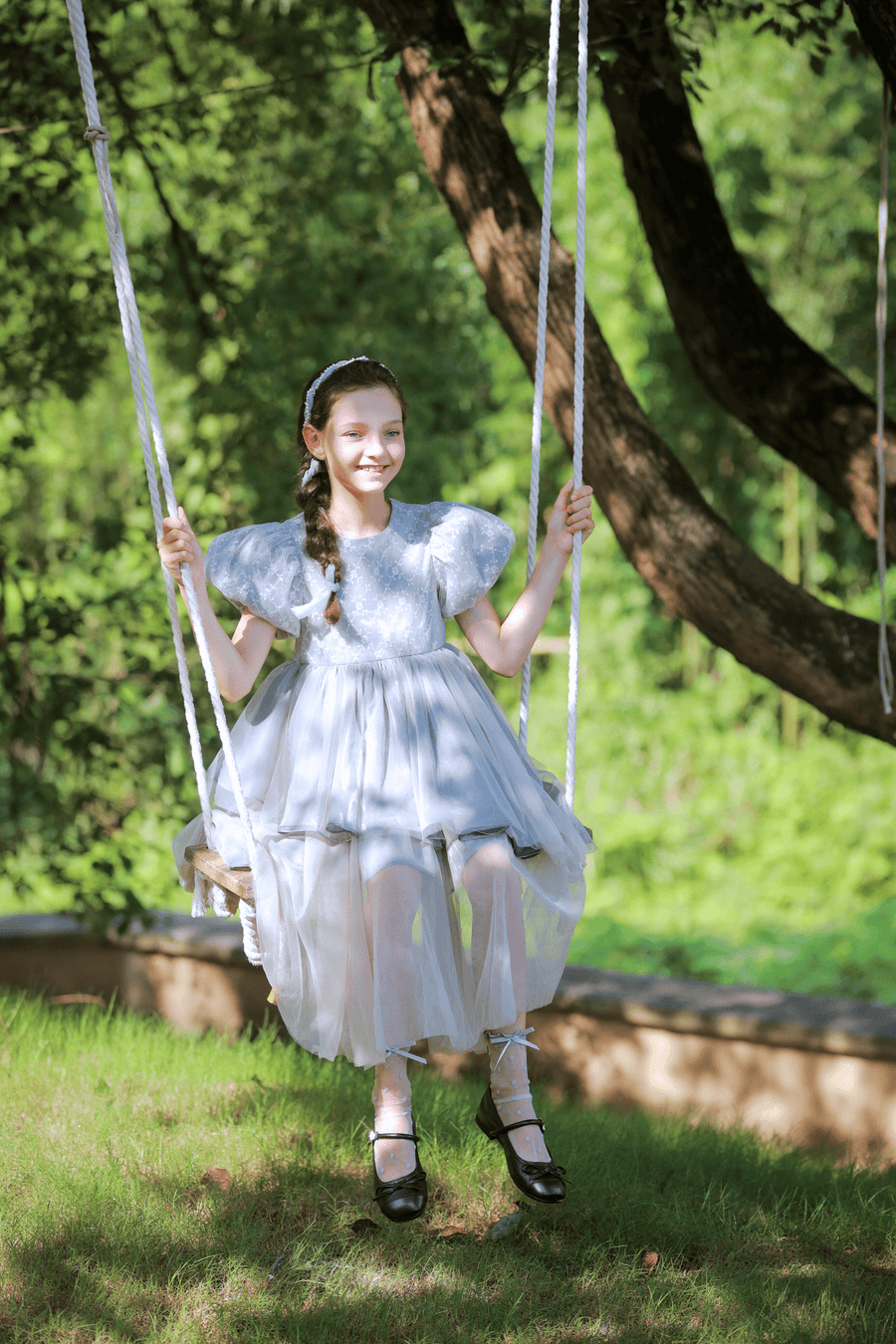 A young girl in the KIKI and JOJO Girls Velvet Asymmetrical Hem Puff Sleeve Dress in gray sits on a swing outdoors, smiling, with green trees and grass in the background.