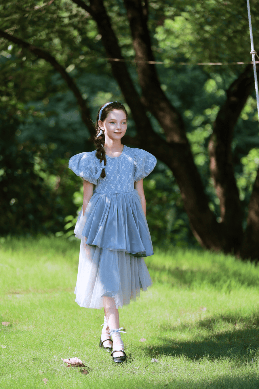 A young girl wears the KIKI and JOJO Girls Velvet Asymmetrical Hem Puff Sleeve Dress in gray as she walks on green grass with trees in the background on a sunny day.