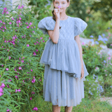 A young girl wears the KIKI and JOJO Girls Velvet Asymmetrical Hem Puff Sleeve Dress in gray, standing in a garden with green plants and blooming flowers, holding a pink flower to her face.