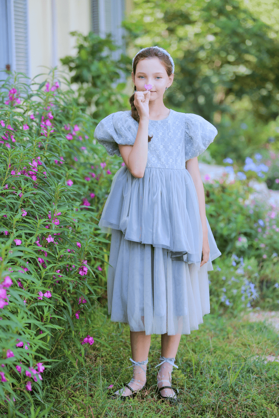 A young girl wears the KIKI and JOJO Girls Velvet Asymmetrical Hem Puff Sleeve Dress in gray, standing in a garden with green plants and blooming flowers, holding a pink flower to her face.