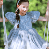 A young girl with braided hair and a headband wears the KIKI and JOJO Girls Velvet Asymmetrical Hem Puff Sleeve Dress in gray while sitting on a swing outdoors.