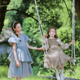Two young girls are outdoors; one stands by a swing while the other, dressed in the KIKI and JOJO Girls Velvet Asymmetrical Hem Puff Sleeve Dress in gray, sits smiling among trees and greenery.