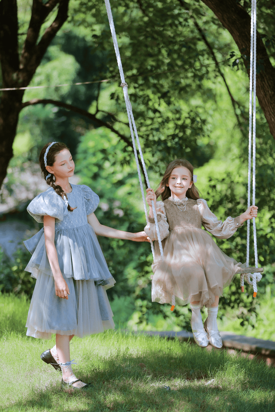 Two young girls are outdoors; one stands by a swing while the other, dressed in the KIKI and JOJO Girls Velvet Asymmetrical Hem Puff Sleeve Dress in gray, sits smiling among trees and greenery.