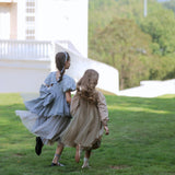 Two young girls wearing KIKI and JOJO Girls Velvet Asymmetrical Hem Puff Sleeve Dress in gray run across a grassy lawn toward a white building, framed by trees and hills.