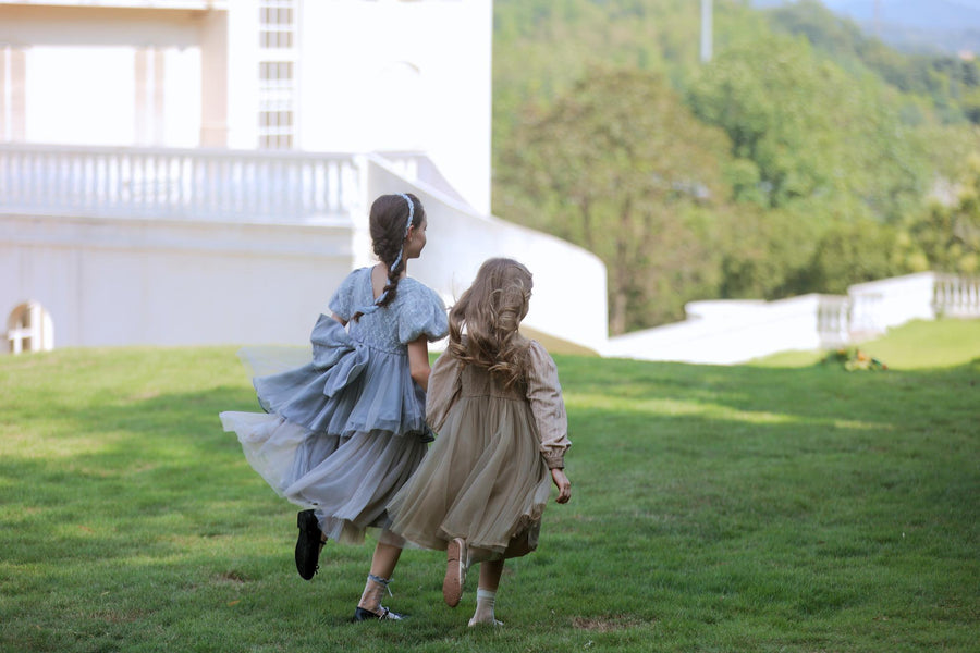Two young girls wearing KIKI and JOJO Girls Velvet Asymmetrical Hem Puff Sleeve Dress in gray run across a grassy lawn toward a white building, framed by trees and hills.