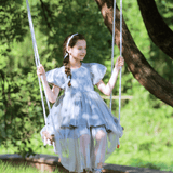 A young girl wearing the KIKI and JOJO Girls Velvet Asymmetrical Hem Puff Sleeve Dress in gray sits on a swing in a sunlit, green outdoor setting.