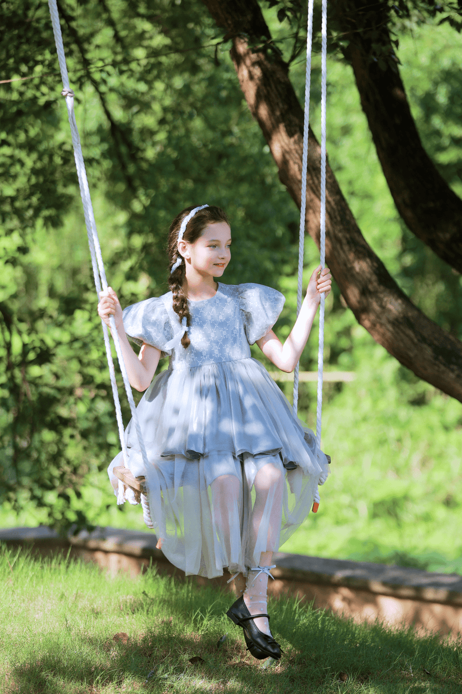 A young girl wearing the KIKI and JOJO Girls Velvet Asymmetrical Hem Puff Sleeve Dress in gray sits on a swing in a sunlit, green outdoor setting.