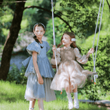 Two young girls, one standing and one sitting on a swing, wear KIKI and JOJO Girls Velvet Asymmetrical Hem Puff Sleeve Dresses in gray, outdoors on grassy ground with trees behind them.