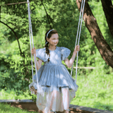 A young girl in a KIKI and JOJO Girls Velvet Asymmetrical Hem Puff Sleeve Dress in gray sits on a swing outdoors, smiling amid green trees and grass on a sunny day.