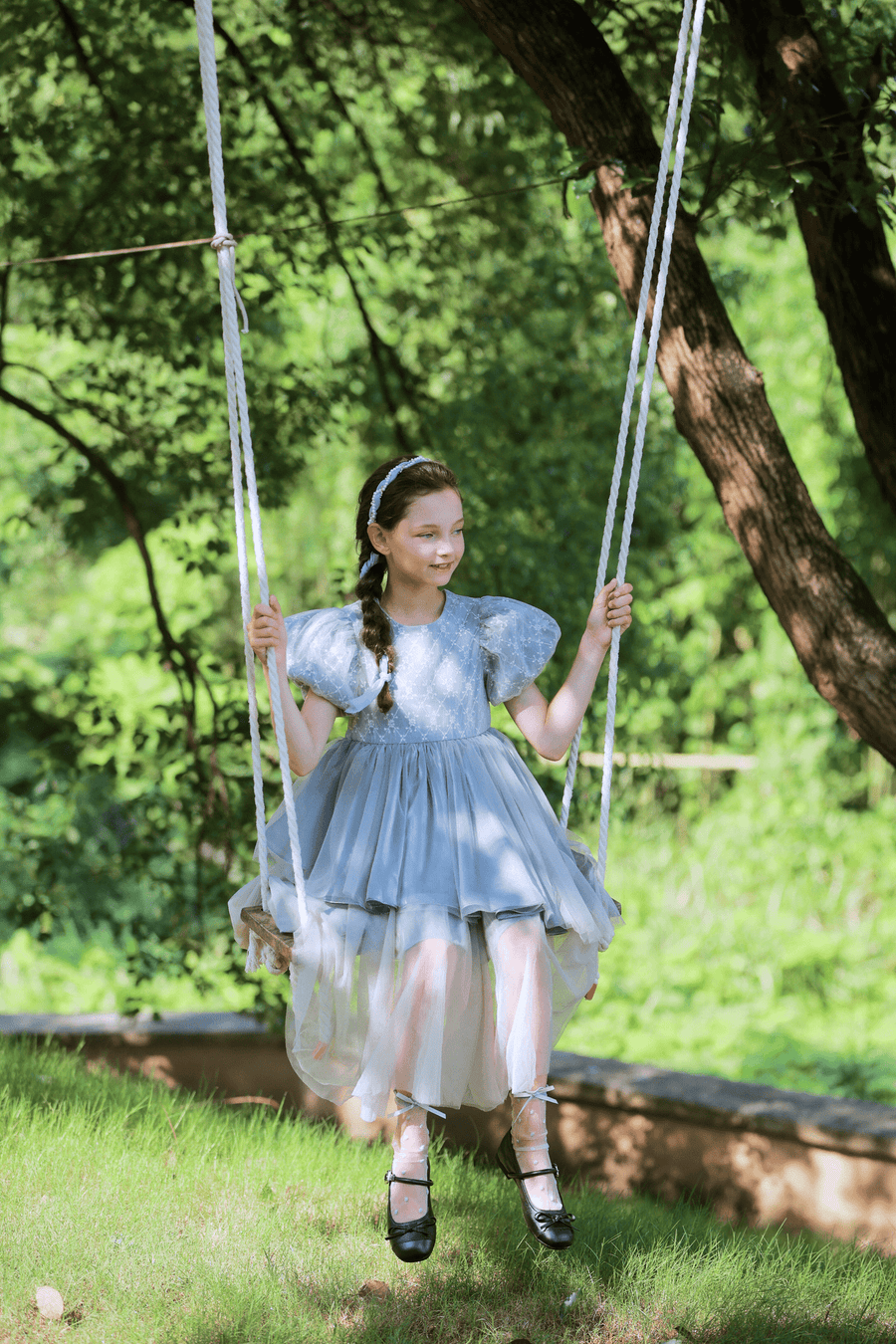 A young girl in a KIKI and JOJO Girls Velvet Asymmetrical Hem Puff Sleeve Dress in gray sits on a swing outdoors, smiling amid green trees and grass on a sunny day.