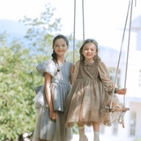 Two young girls in pastel dresses pose outdoors; one stands on the grass, while the other, in a KIKI and JOJO Girls Velvet Asymmetrical Hem Puff Sleeve Dress - Gray, sits on a rope swing among trees with a building behind them.