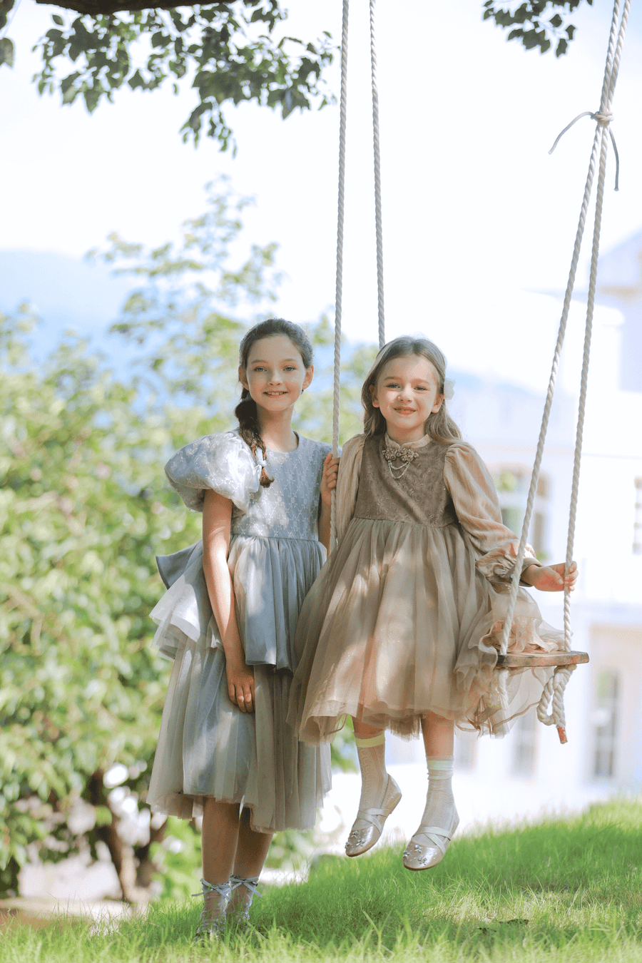 Two young girls in pastel dresses pose outdoors; one stands on the grass, while the other, in a KIKI and JOJO Girls Velvet Asymmetrical Hem Puff Sleeve Dress - Gray, sits on a rope swing among trees with a building behind them.