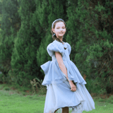 A young girl in a gray Girls Velvet Asymmetrical Hem Puff Sleeve Dress by KIKI and JOJO stands on grass, framed by green trees in the background.