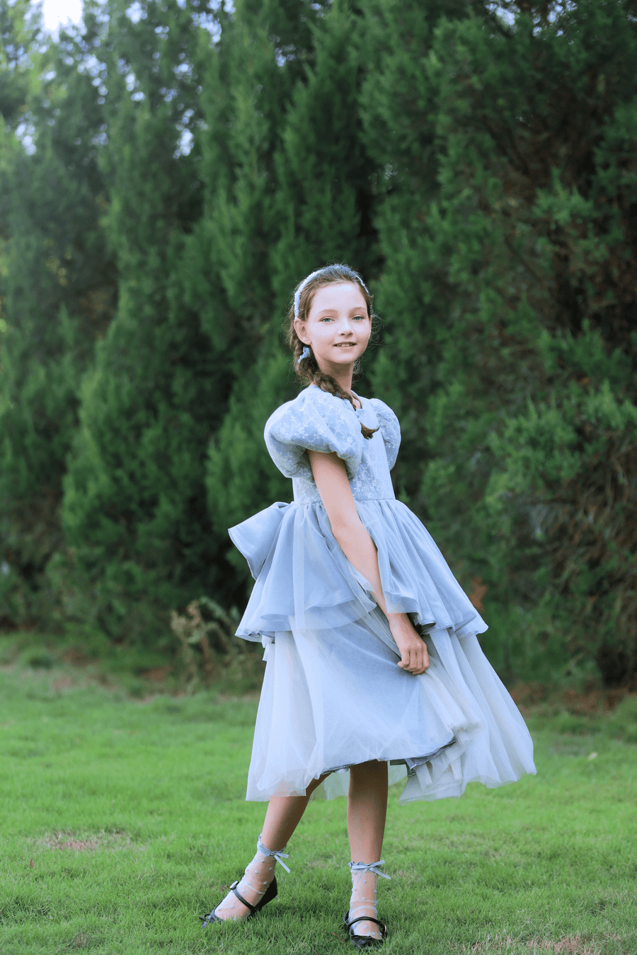 A young girl in a gray Girls Velvet Asymmetrical Hem Puff Sleeve Dress by KIKI and JOJO stands on grass, framed by green trees in the background.