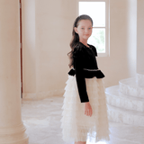 A young girl poses indoors by tall pillars and a window, wearing the KIKI and JOJO Girls Velvet & Tulle Sparkle Dress in Black/Ivory, paired with black shoes and a headband for an elegant, charming look.