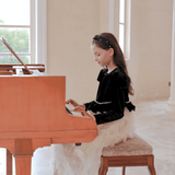 A young girl in a KIKI and JOJO Girls Velvet & Tulle Sparkle Dress - Black/Ivory sits on a padded bench, playing a wooden piano in a bright room with large windows.