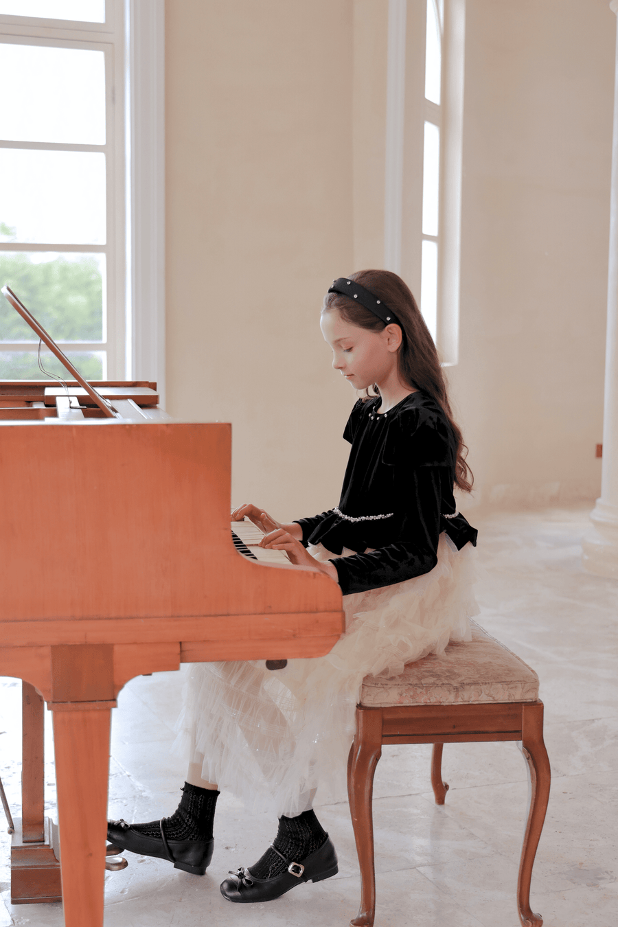 A young girl in a KIKI and JOJO Girls Velvet & Tulle Sparkle Dress - Black/Ivory sits on a padded bench, playing a wooden piano in a bright room with large windows.