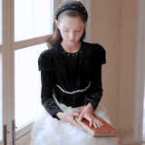 A young girl sits by a window wearing the KIKI and JOJO Girls Velvet & Tulle Sparkle Dress in Black/Ivory, her hands resting on a closed book in her lap.