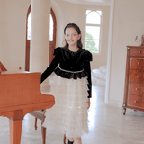 A young girl wearing the KIKI and JOJO Girls Velvet & Tulle Sparkle Dress in Black/Ivory stands beside a wooden piano in a spacious, well-lit room with marble floors.