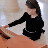 A young girl in a KIKI and JOJO Girls Velvet & Tulle Sparkle Dress - Black/Ivory sits at a wooden piano, gazing down at the keys as she prepares to play.