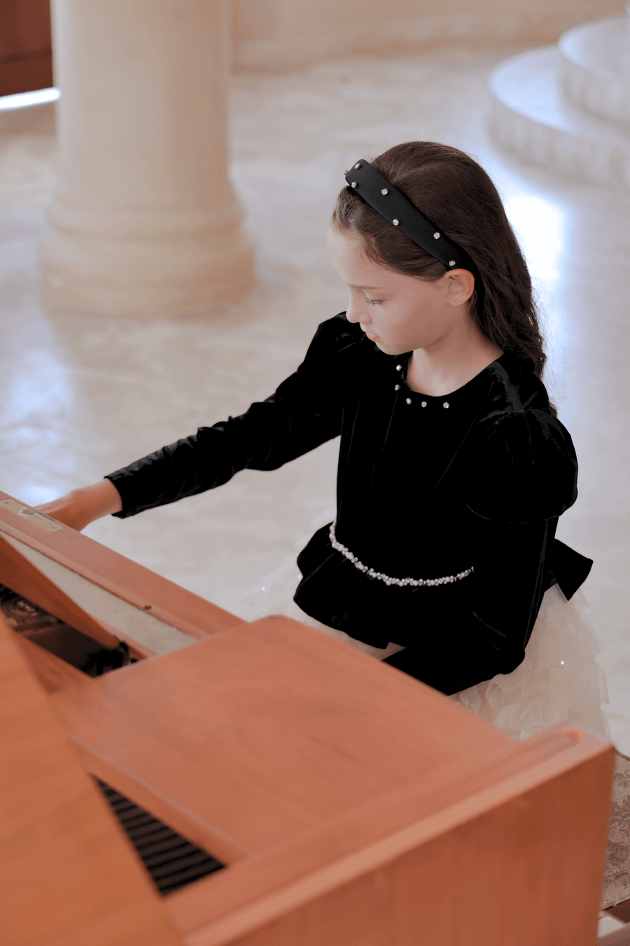 A young girl in a KIKI and JOJO Girls Velvet & Tulle Sparkle Dress - Black/Ivory sits at a wooden piano, gazing down at the keys as she prepares to play.