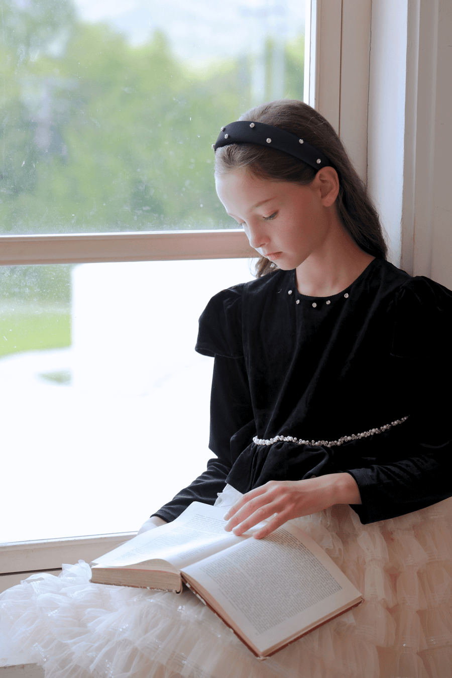 A girl in the KIKI and JOJO Girls Velvet & Tulle Sparkle Dress - Black/Ivory sits by a window, pearl headband in her hair, reading an open book as gentle daylight fills the room.