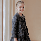 A young girl with light hair in a bun smiles at the camera, wearing the KIKI and JOJO Girls Vintage Beaded Tweed Jacket in gray—an elegant, vintage-inspired look beside a light curtain and beige wall.