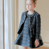 A young girl with light hair in a bun exudes vintage elegance, wearing the KIKI and JOJO Girls Vintage Beaded Tweed Jacket in gray as she stands indoors by a curtain.