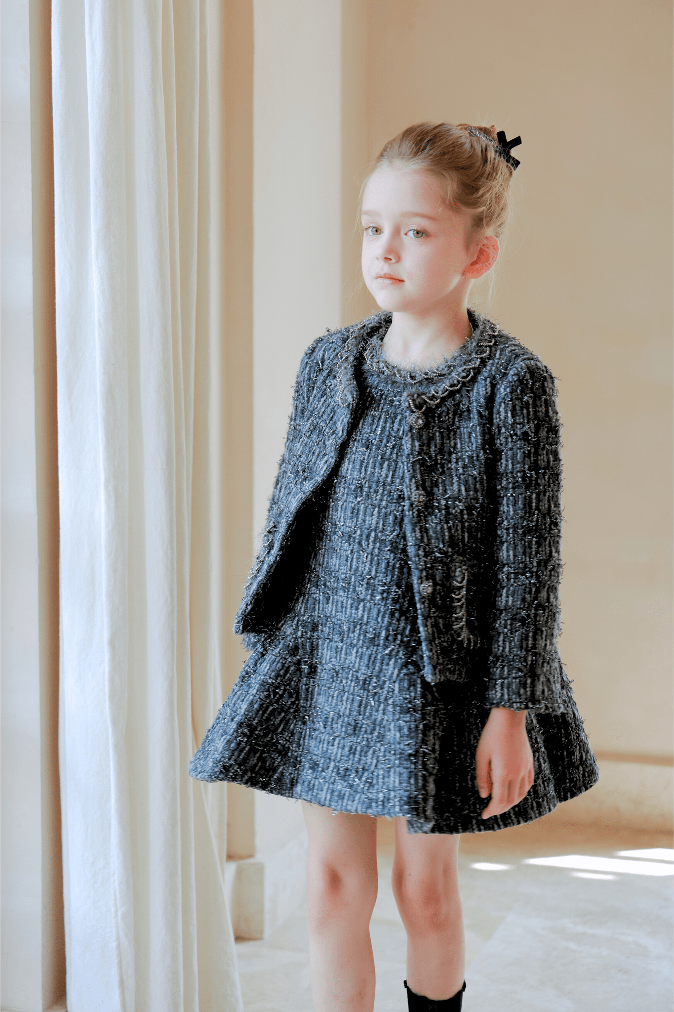 A young girl with light hair in a bun exudes vintage elegance, wearing the KIKI and JOJO Girls Vintage Beaded Tweed Jacket in gray as she stands indoors by a curtain.