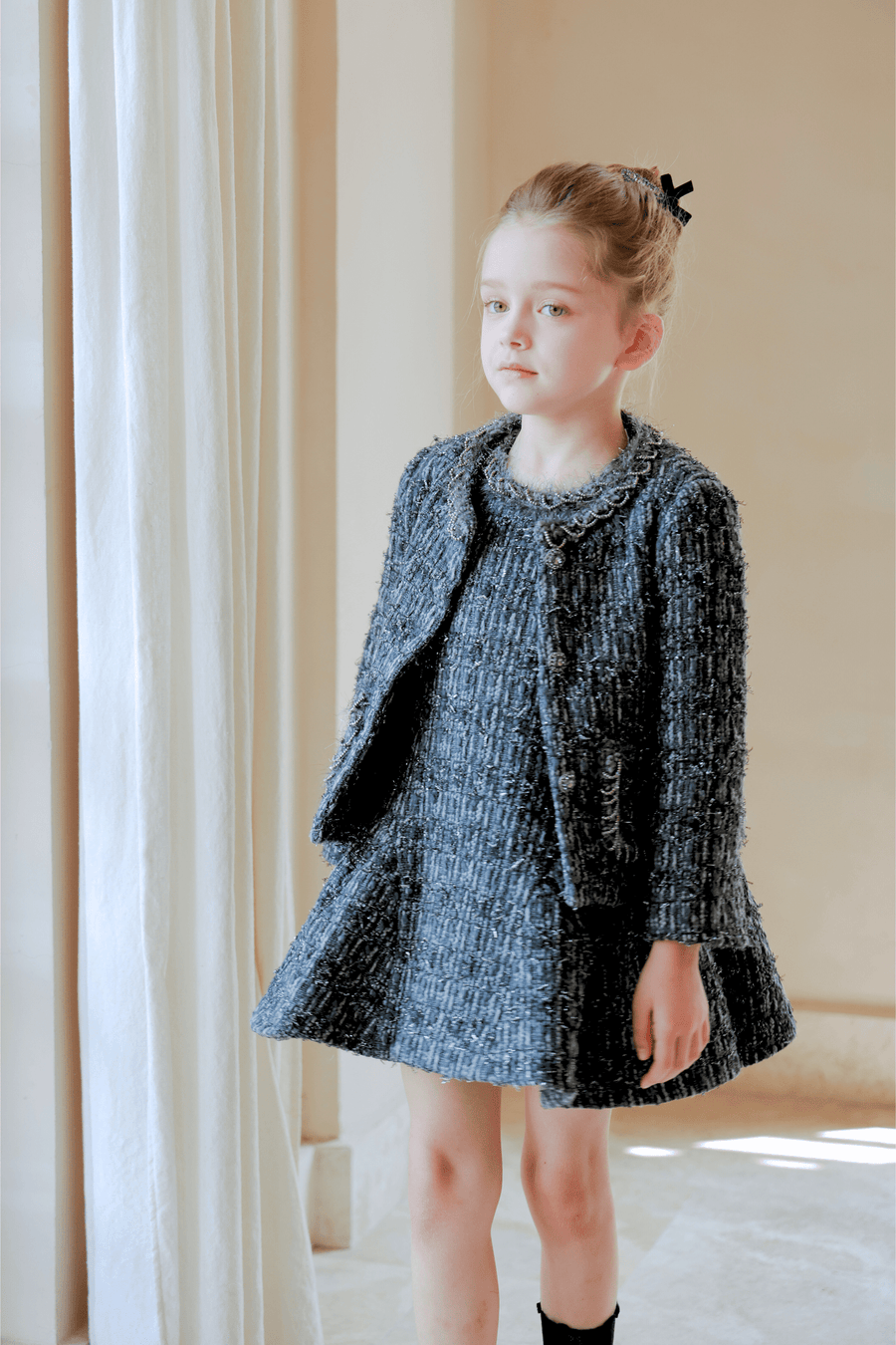 A young girl with light hair in a bun exudes vintage elegance, wearing the KIKI and JOJO Girls Vintage Beaded Tweed Jacket in gray as she stands indoors by a curtain.
