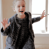 A young girl smiles indoors, raising her hands in natural light, wearing the KIKI and JOJO Girls Vintage Beaded Tweed Jacket in gray, exuding vintage-inspired elegance.