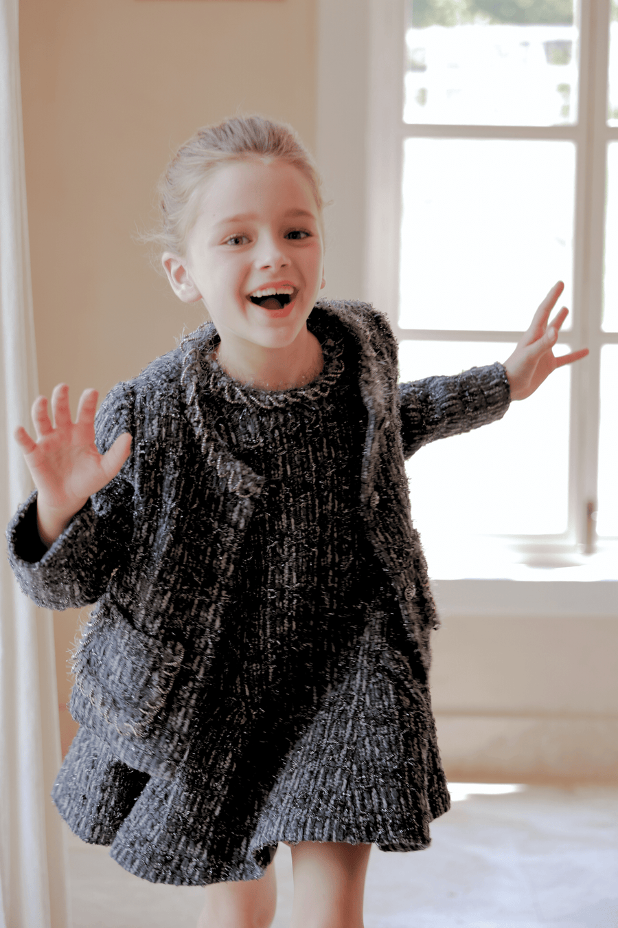 A young girl smiles indoors, raising her hands in natural light, wearing the KIKI and JOJO Girls Vintage Beaded Tweed Jacket in gray, exuding vintage-inspired elegance.