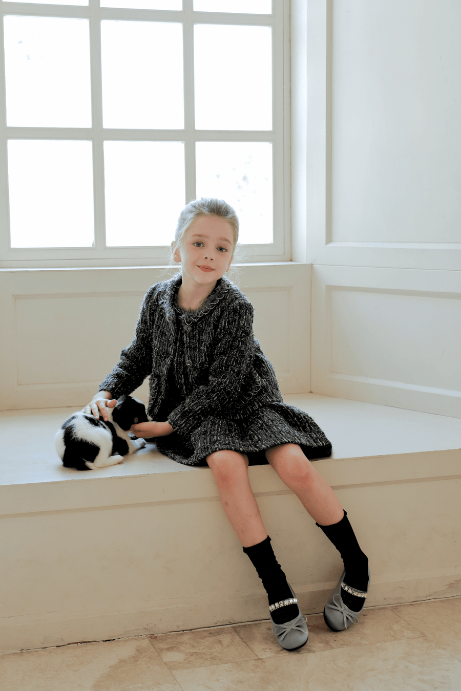 A young girl wearing the KIKI and JOJO Girls Vintage Beaded Tweed Jacket in gray sits by a window, gently petting a black and white cat on her lap.