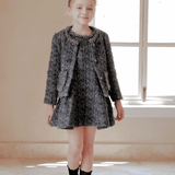 A young girl stands indoors in the KIKI and JOJO Girls Vintage Beaded Tweed Jacket - Gray, exuding classic elegance as soft window light highlights her sophisticated children's outerwear.
